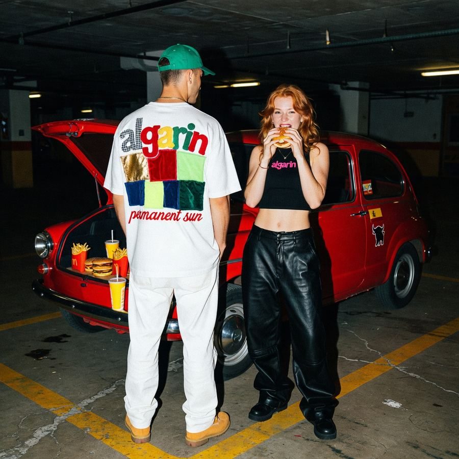 Pareja con ropa de una marca de streetwear junto a un coche rojo en un aparcamiento, imagen de campaña